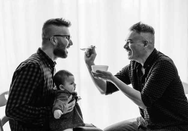 Male gay couple with adopted baby girl at home Two handsome dads feed the baby girl on kitchen Male babysitters Lgbt family at home Diversity concept