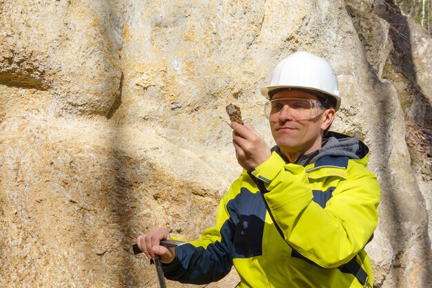 Male geologist in helmet and protective glasses examines a sample of the mineral outdoors against a rock