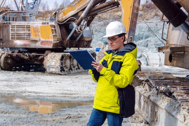 Un geologo maschio o un ingegnere minerario scrive qualcosa in una custodia per mappe in mezzo a una cava con attrezzature da costruzione