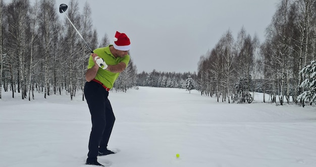 Male golfer in santa claus hat in winter on golf course with golf clubs hitting ball