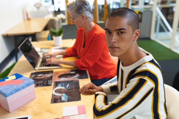 Male graphic designer looking at camera while coworker using laptop in the background