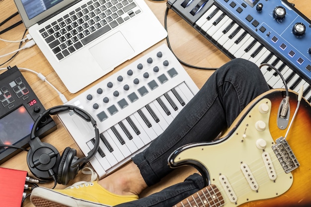 Фото Гитарист мужского пола с электрогитарой в современной домашней студии или репетиционной комнате. молодой человек производит музыку с электронными процессорами эффектов, синтезатором и ноутбуком