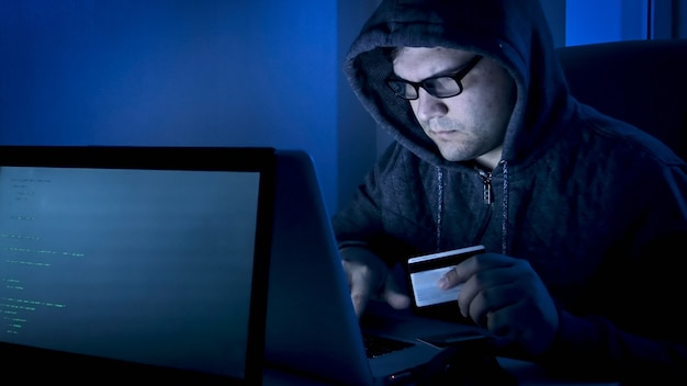 Male hacker in hood holding credit card and stealing money from online banking system