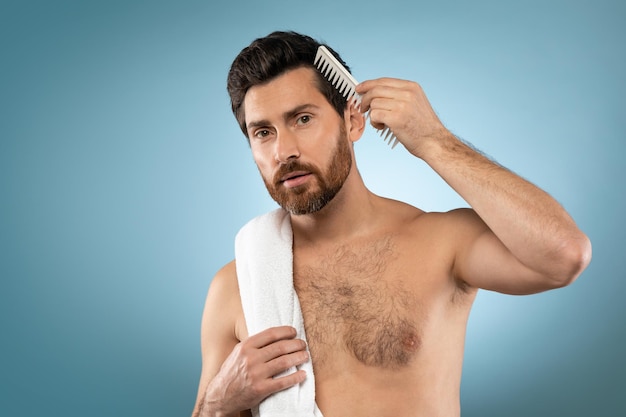 Male haircare Handsome shirtless man brushing hair with hairbrush standing with towel on shoulder blue background