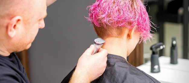 Male hairdresser shaves neck of a young caucasian woman with a short pink hairstyle by electric shaver in a hairdresser salon close up