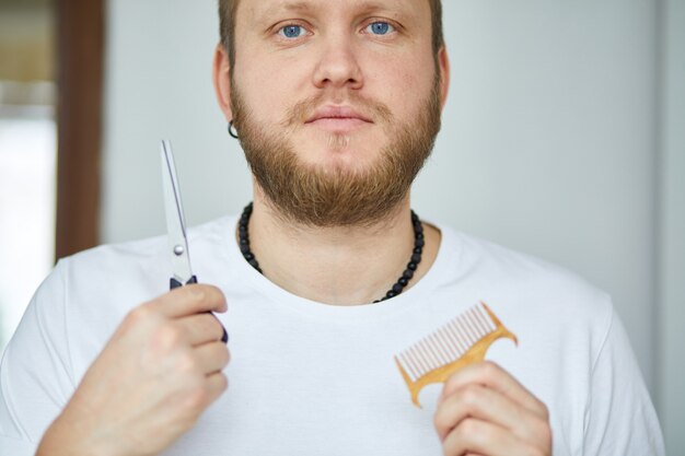 A male hairdresser wih bread holds scissors and a comb in his hands, barbershop