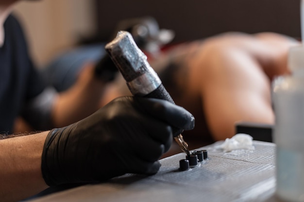 Male hand in a black glove with a tattoo machine takes ink and makes a tattoo in the studio. Man professional tattoo artist in work process, close-up