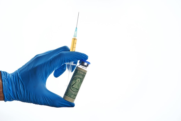 Male hand in a blue glove holds vial dose of COVID-19 vaccine with syringe wrapped dollar bill against white backdrop, global vaccination and money abuse concept