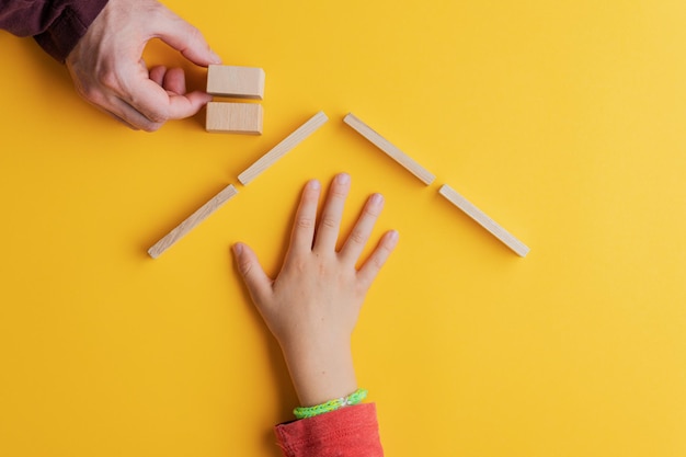 Mano maschile che costruisce una casa di pioli di legno e cubi per una mano di bambino che vi si ripari. su sfondo giallo.