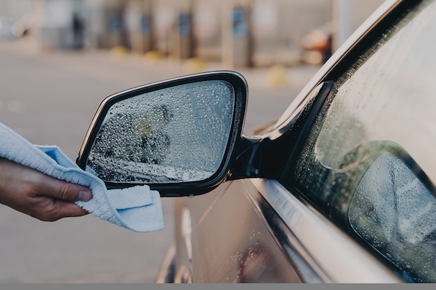 Male hand cleaning car outdoors with rag wiping water with microfiber cloth on side rearview mirror