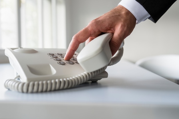 Foto mano maschile che compone un numero di telefono per effettuare una telefonata su un classico telefono fisso bianco su una scrivania in ufficio.