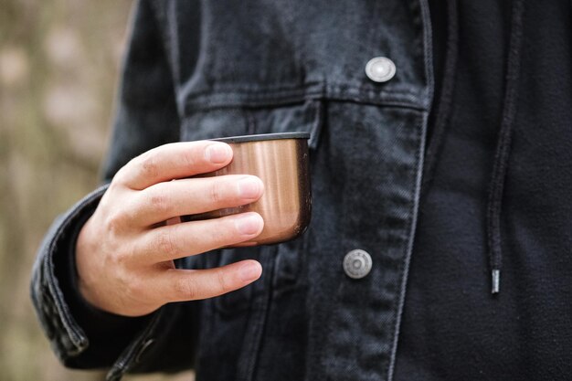 Foto male hand holding a cup of thermos with hot tea on a background of pine forest drink hot tea or coffee in the forest thermo mug with a drink in hand