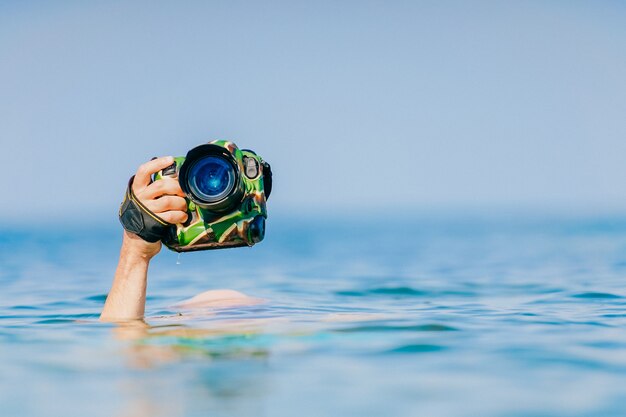 Male hand holding photocamera above water in sea.