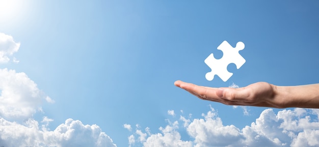 Male hand holding puzzle icon on blue background. pieces representing the merging of two companies or joint venture, partnership, Mergers and acquisition concept.