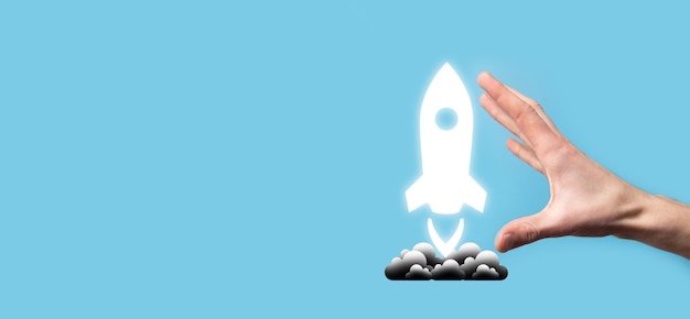 Male hand holding rocket icon that takes off launch on blue
background rocket is launching and flying out business start up
icon marketing on modern virtual interfacestart up concept