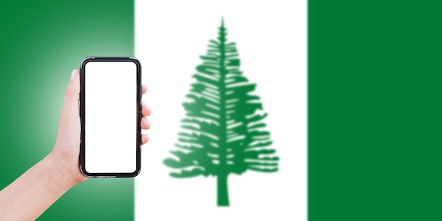 Male hand holding smartphone with blank on screen on background of blurred flag of Norfolk Island Closeup view