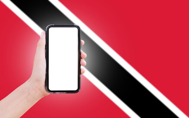 Male hand holding smartphone with blank on screen on background of blurred flag of Trinidad and Tobago Closeup view