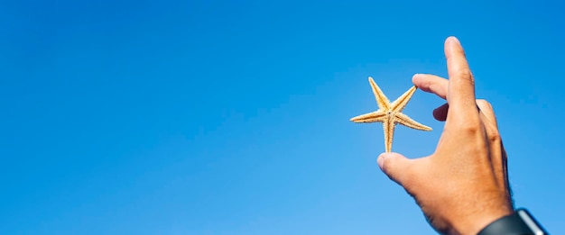 Male hand holding a starfish on a blue sky background Banner