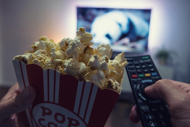 Male hand holding TV remote control and popcorn box Point of view shot