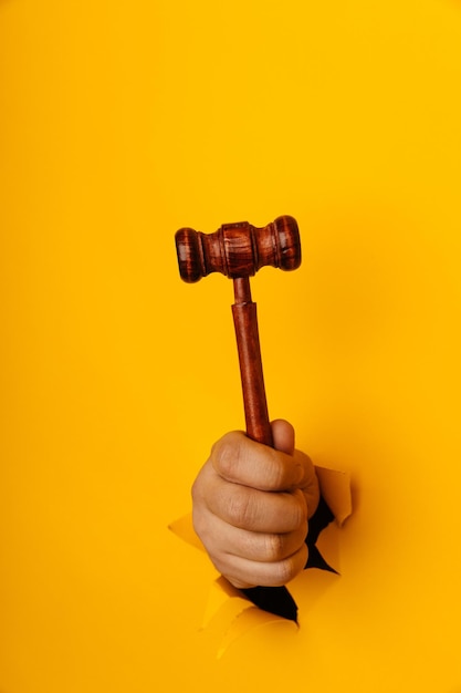 Male hand holding a wooden gavel through torn yellow background Law and auction aconcept Vertical image