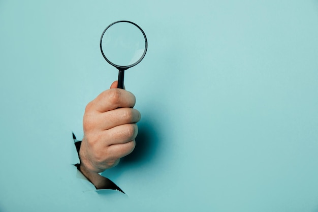 Male hand holds black magnifying glass in torn hole of blue\
background searching information concept