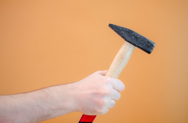 Male hand holds a hammer with a wooden handle on an orange background. Close-up