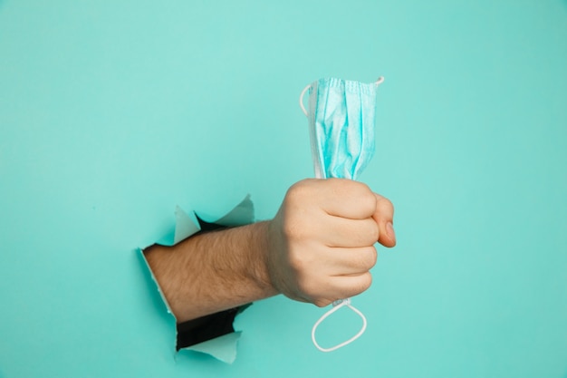 A male hand holds a medical mask through a hole in a blue wall. Medical breakthrough concept.