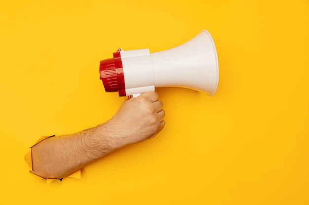 Male hand holds a megaphone from a hole in the wall on a yellow background Advertising concept