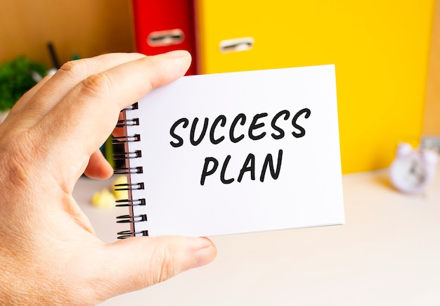 한 남성의 손이 SUCCESS PLAN 텍스트가 있는 봄철 메모장을 들고 있습니다. 사무 공간. 비즈니스 개념입니다.