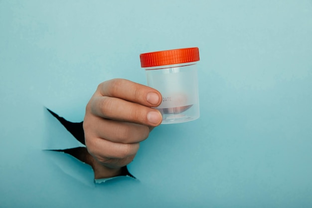 Photo male hand holds a plastic jar for urine tests the hand is sticking out of a torn hole in a blue background