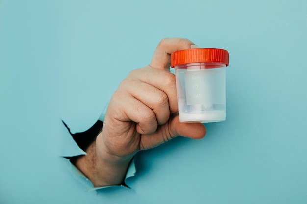 Male hand holds a plastic jar for urine tests the hand is sticking out of a torn hole in a blue background