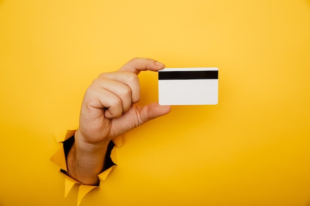 Foto la mano maschile tiene la carta di credito in un buco strappato di sfondo giallo shopping online acquisto di prodotti concetto