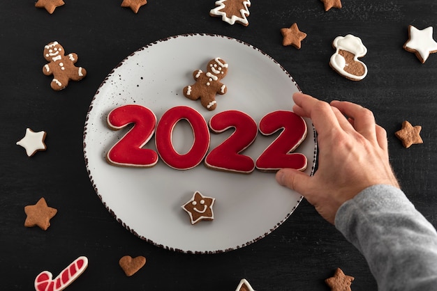 Male hand lays down tasty glazed painted cookies on white plate: numbers 2022, stars and fir-tree. New Year food concept