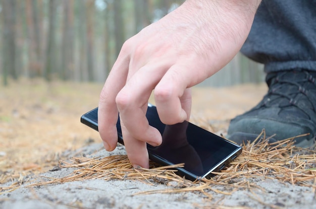 Male hand picking up lost mobile phone from a ground in autumn fir wood path The concept of finding a valuable thing and good luck