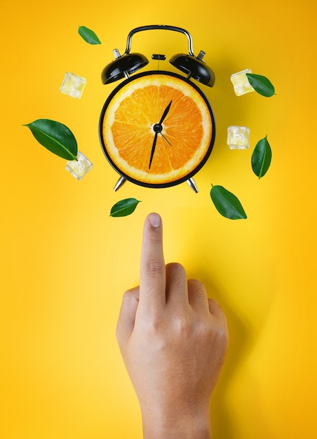 Male hand pointing alarm clock of orange fruit green leaves and ice cube flying around