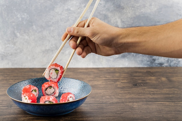 Male hand taking Philadelphia roll with chopsticks from plate . 