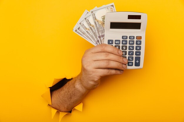 Foto mano maschile con calcolatrice e banconote in dollari attraverso un foro di carta in primo piano sfondo giallo