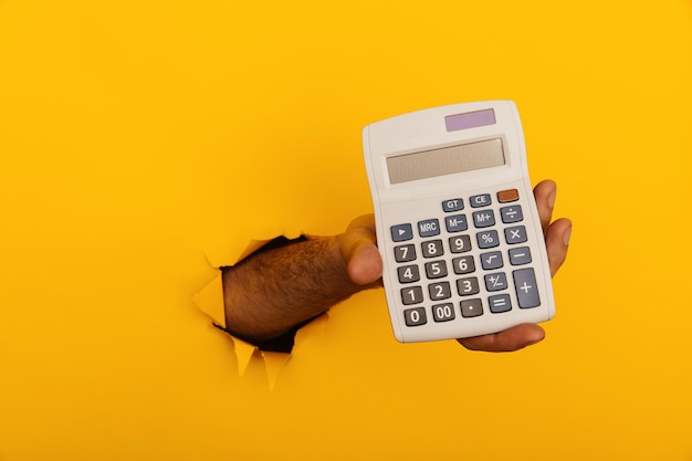 Male hand with calculator through a paper hole in yellow background with closeup