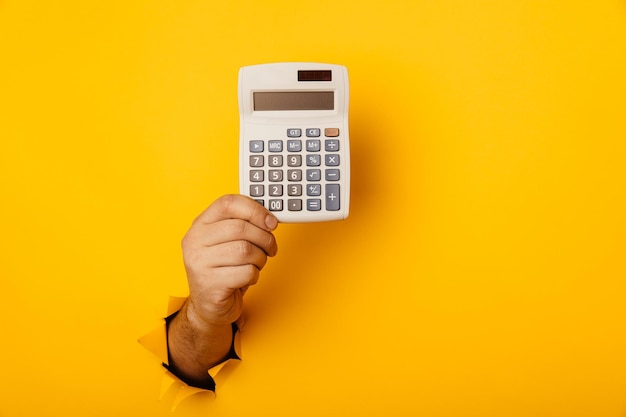 Photo male hand with calculator through a paper hole in yellow background with copy space