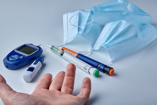 Male hand with a drop of blood on a finger, a glucometer, insulin syringe pens and medical masks on a white table. Diabetes and coronavirus