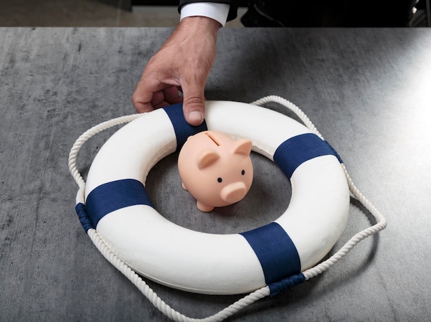 Male hand with little piggy bank inside of lifebuoy safe money
blue and white life buoy on wooden desk table background assets
wealth money saving and security by insurance concept copy
space