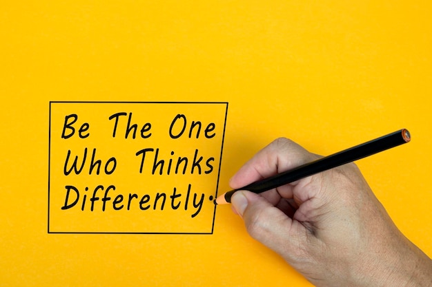 Male hand writes in black pencil the word be the one who things differently on a yellow background encouragement words