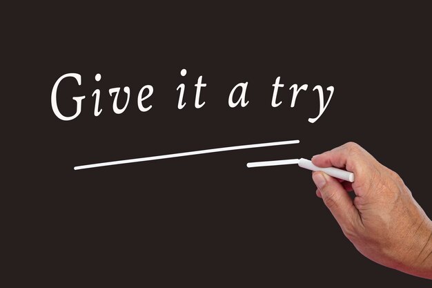 Male hand writes in white chalk pencil the word Give It A Try on a black background Encouragement phrases and business concept