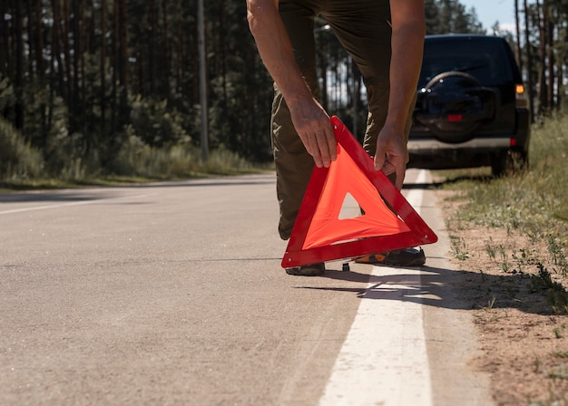 写真 夏に壊れた車の近くの道路側に赤い三角形の警告サインを置く男性の手のクローズアップ