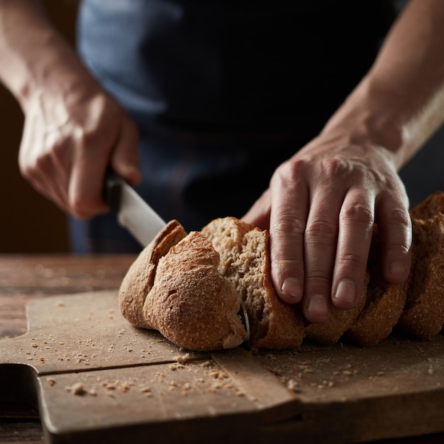 Male hands cutting wheaten bread on the wooden board. Close up. The healthy eating and traditional bakery concept