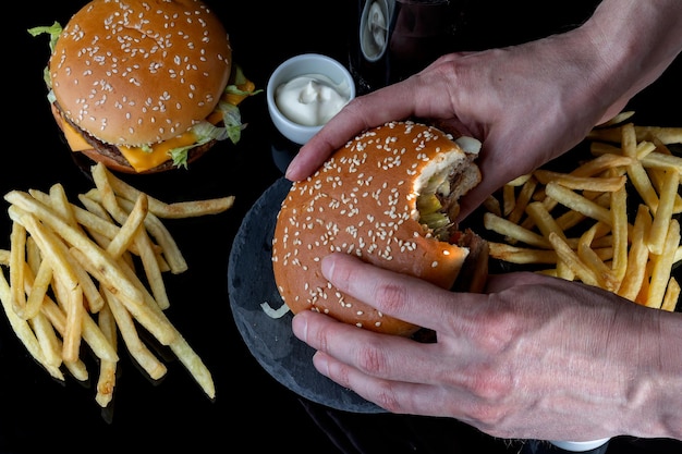 Male hands firmly hold a bitten hamburger fresh tasty hamburger french fries cola on black glass