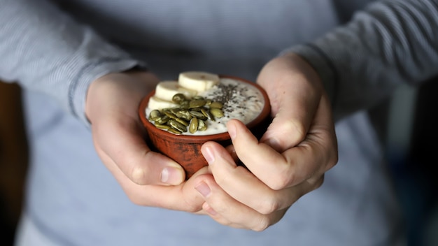 Male hands hold a bowl of yogurt. A man with a spoon eats yogurt with seeds. Chia pudding with pumpkin seeds and banana.