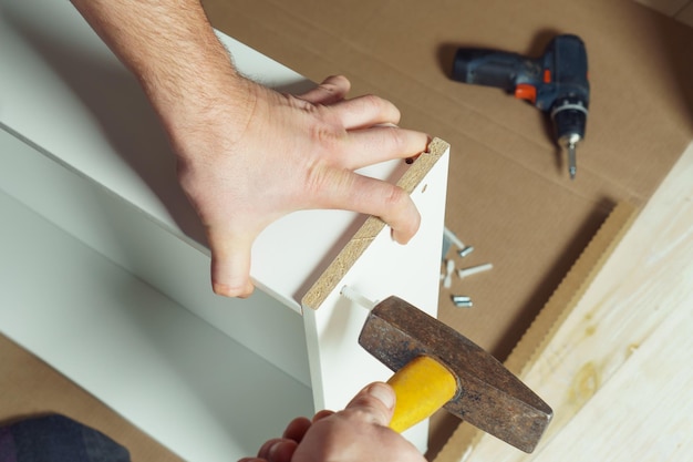 Le mani maschili tengono il martello di metallo con la maniglia di plastica gialla che guida il tassello che assembla il cassetto bianco del comò o dell'armadio