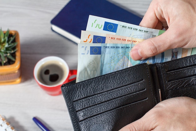 ユーロ紙幣でいっぱいの黒い革の財布を持っている男性の手背景にコーヒーノートブックのカップ