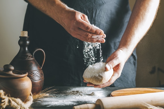 Мужские руки насыпают муку на тесто рядом с глиняным горшком, бутылку с маслом и скалку на темном столе, пока готовят